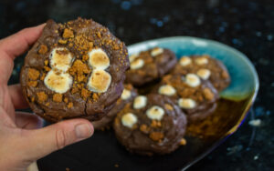 Hand holding up a S'mores Brownie Cookie. More cookies are shown on a plate in the background.