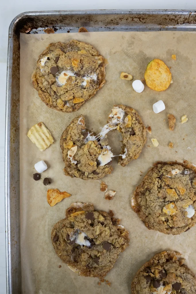 Kitchen sink cookies on a baking sheet, with a marshmallow stretching between two broken halves of a cookie.