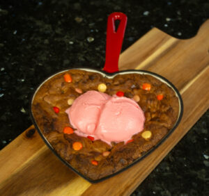 Chocolate Chip Skillet Cookie for Two, in a red heart-shaped skillet and topped with two scoops of Cinnamon Hearts-flavoured ice cream.