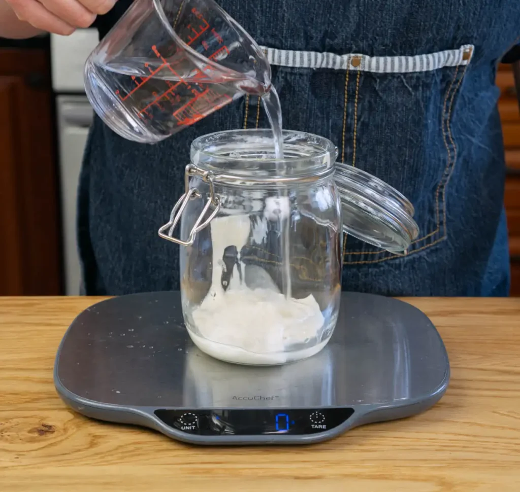 Feeding sourdough starter. Pouring water into a glass jar of sourdough starter on a scale.