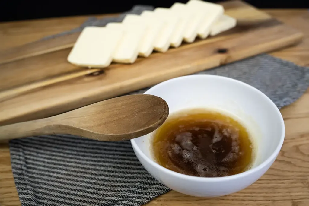 A bowl of brown butter with a wooden spoon, and slices of butter on a wooden board in the background.