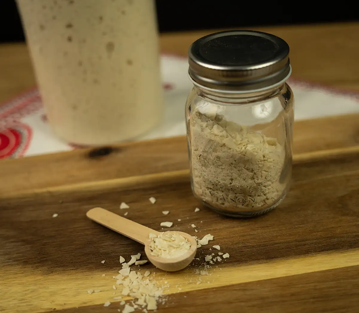 Small mason jar filled with flakes of dehydrated sourdough starter, with a small wooden spoon full of starter flakes in the foreground, and a large mason jar full of active sourdough starter in the background.