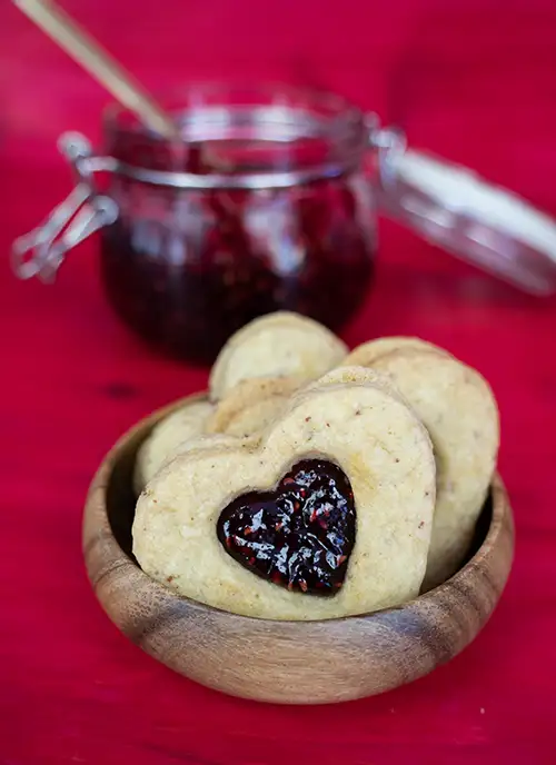 A small bowl with three heart-shaped cookies filled with raspberry jam, and an open jar of jam in the background.
