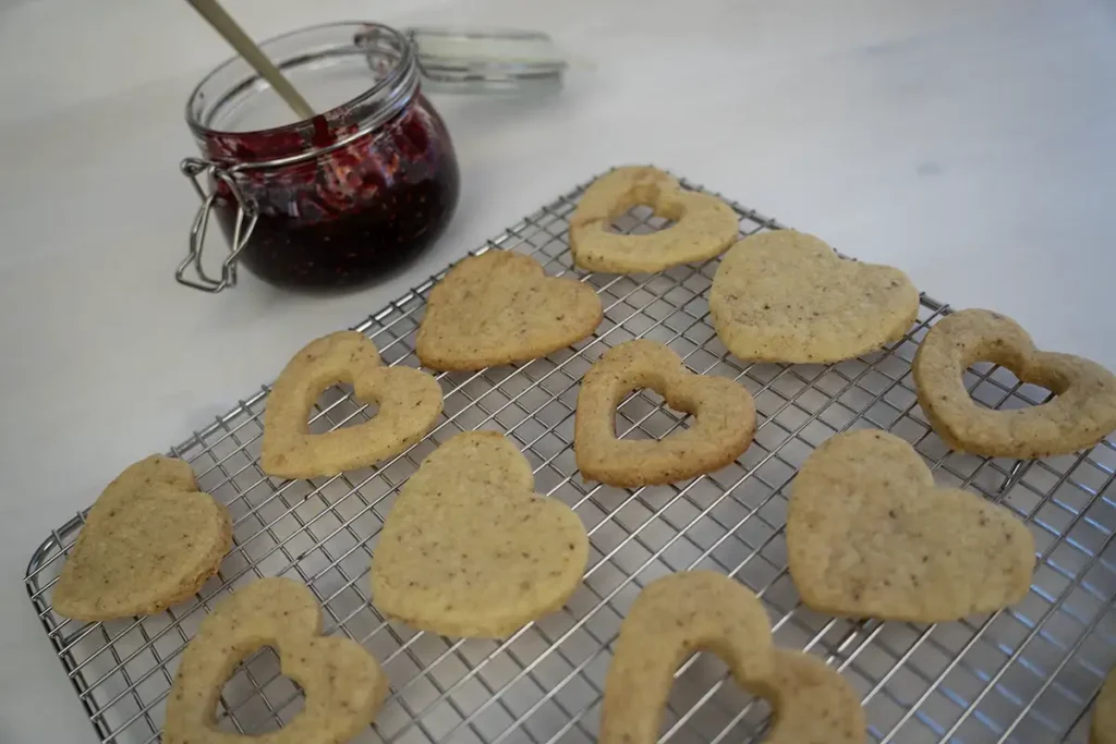 Wire cooling rack with heart-shaped sourdough cut-out cookies and a jar of raspberry jam in the background.
