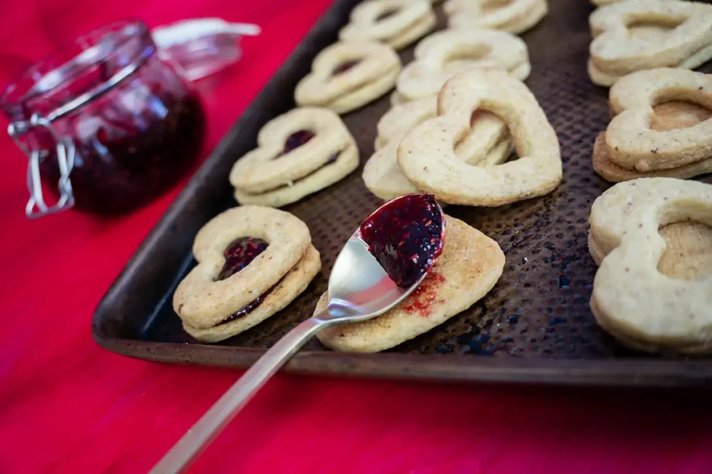 A baking sheet with heart-shaped cookies being filled with raspberry jam.