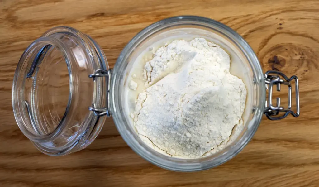 Top view of an open glass jar with sourdough starter being fed with flour and water.