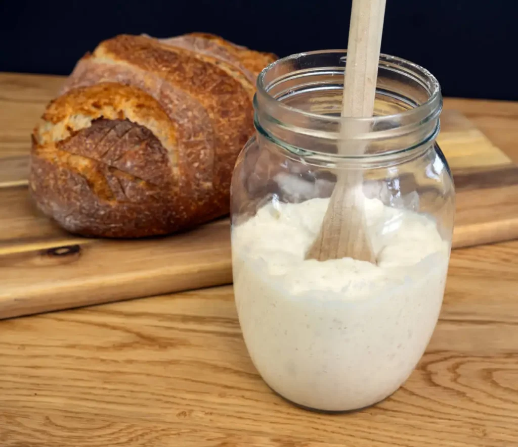 Open mason jar full of sourdough starter with a wooden spoon, and a loaf of sourdough bread in the background.