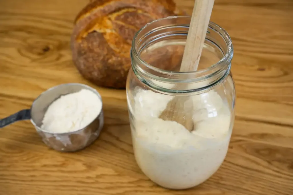 A glass mason jar half-full of bubbly sourdough starter and a wooden spoon standing up in it. In the background is a loaf of sourdough bread and a measuring cup with flour.