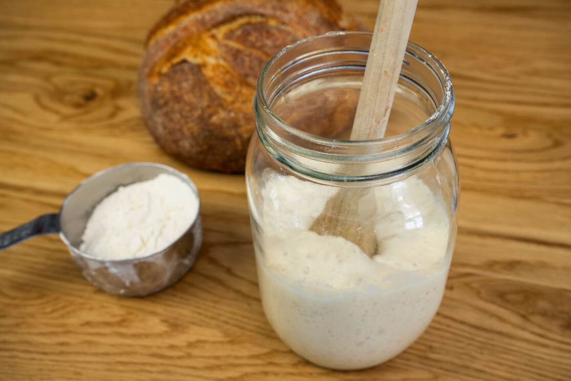 Jar of sourdough starter with a wooden spoon, a measuring cup with flour, and a loaf of sourdough bread in the background.