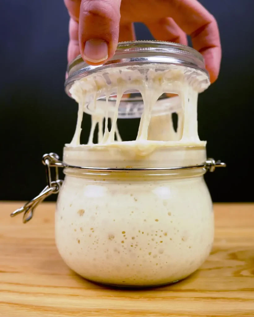 Lifting the lid off a jar of sourdough starter, stretching strands of starter between the jar and the lid.