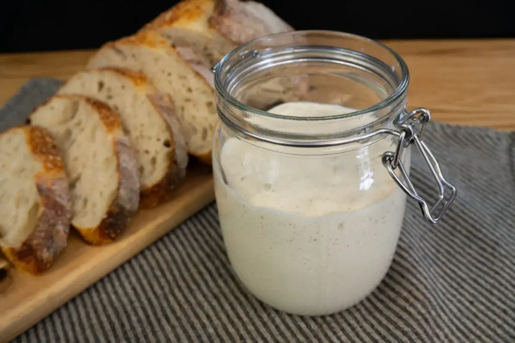 Open jar of sourdough starter with a loaf of sliced sourdough bread in the background.