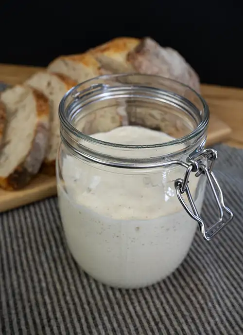 A glass jar of sourdough starter, with a loaf of sourdough bread in the background.