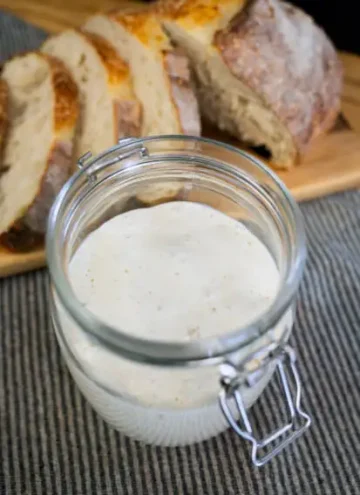 Open jar of bubbly sourdough starter, with a loaf of sliced sourdough bread in the background.