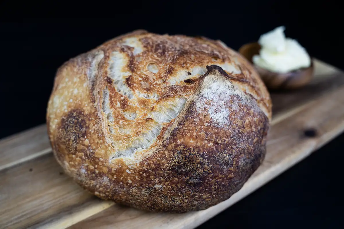 Loaf of sourdough bread on a board of wood, with a dish of butter in the background.