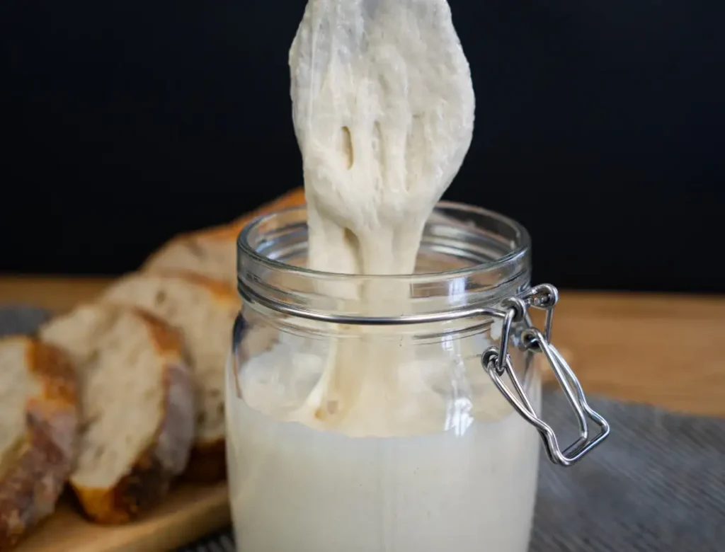 Wooden spoon pulling sourdough starter out of a glass jar, with a loaf of sliced sourdough bread in the background.