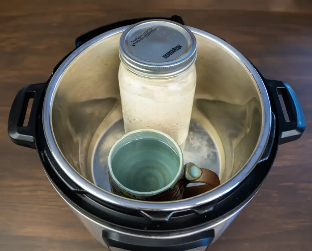 A jar of sourdough starter  with a mug of hot water in an Instant Pot.