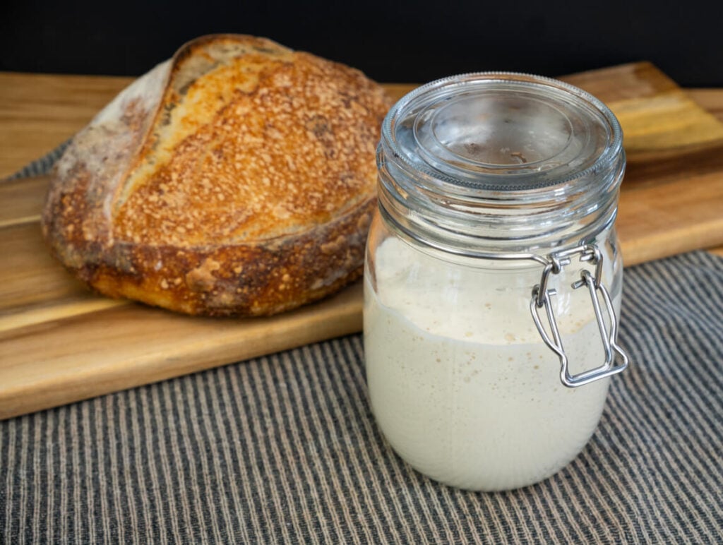 Jar of sourdough starter with a loaf of sourdough bread in the background.
