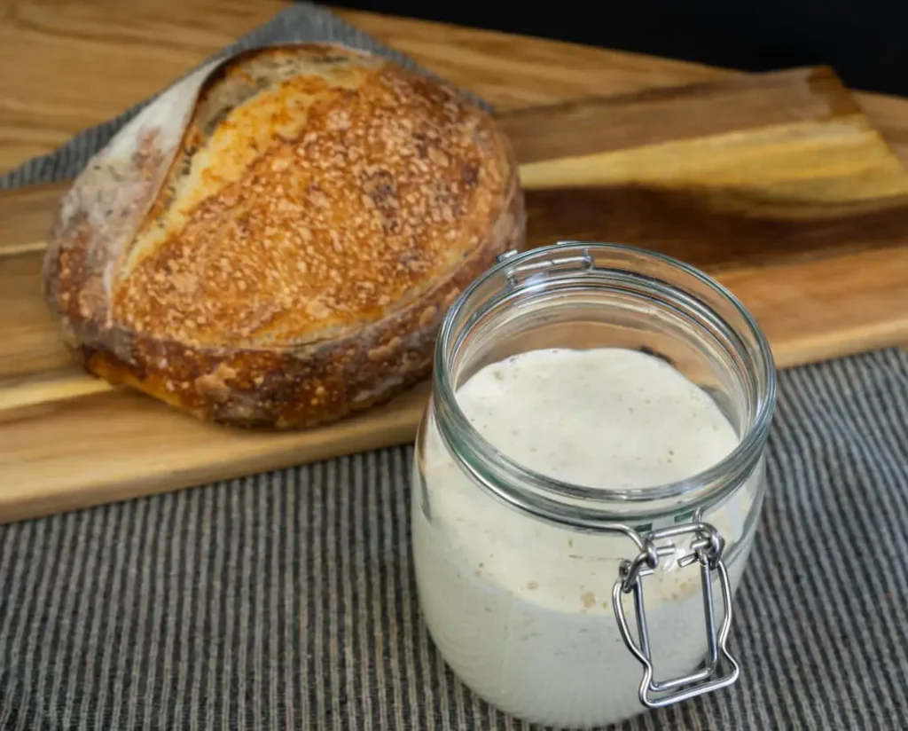 Jar of sourdough starter, with a loaf of sourdough bread in the background.