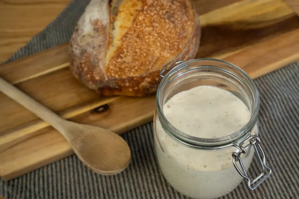 Open jar of sourdough starter with a wooden spoon and a loaf of sourdough bread in the background.