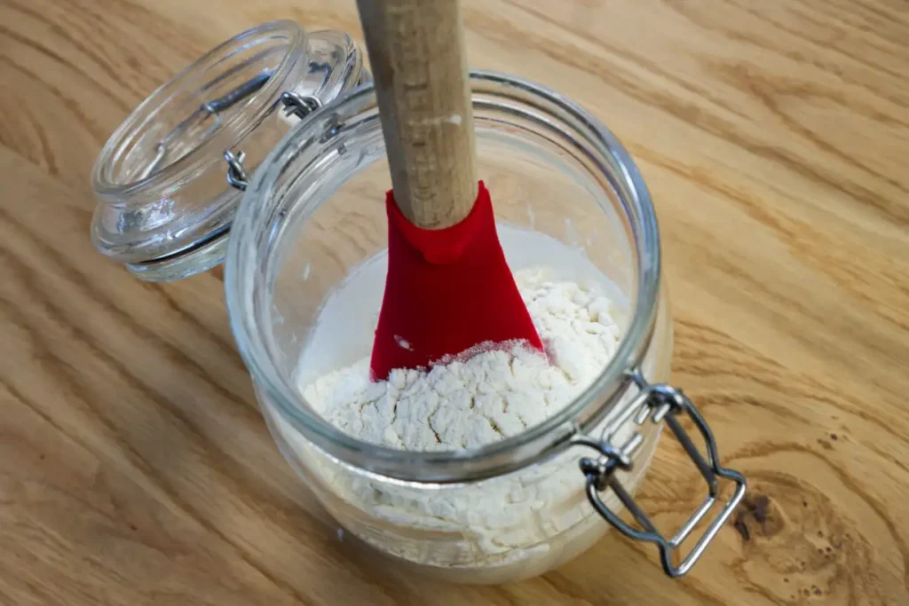 A rubber spatula in a glass jar with sourdough starter, water, and flour. Feeding sourdough starter.