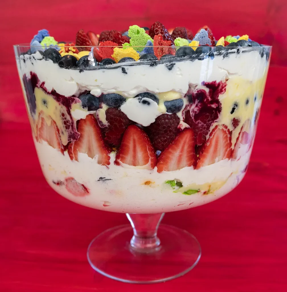 Side view of a fresh fruit trifle with sliced strawberries, blueberries, whipped cream, raspberries, lemon curd, and macaron crumbles.