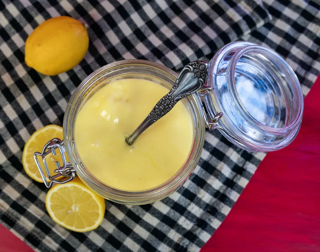 Top view of an open jar of lemon curd, with fresh cut lemons in the background.