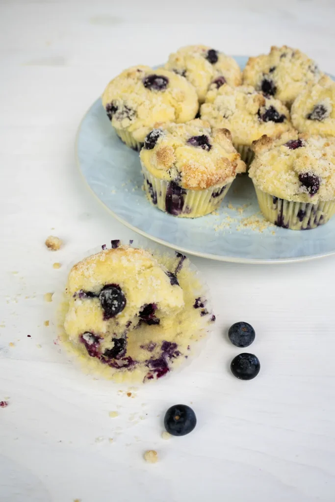 Plate of sourdough blueberry lemon muffins. One muffin is in the foreground with an open paper wrapping, and a couple of loose blueberries next to it.