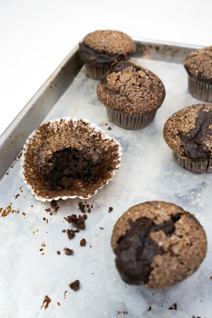 Sourdough discard double chocolate banana muffins scattered on a baking sheet. One muffin has had a bite taken out of it.