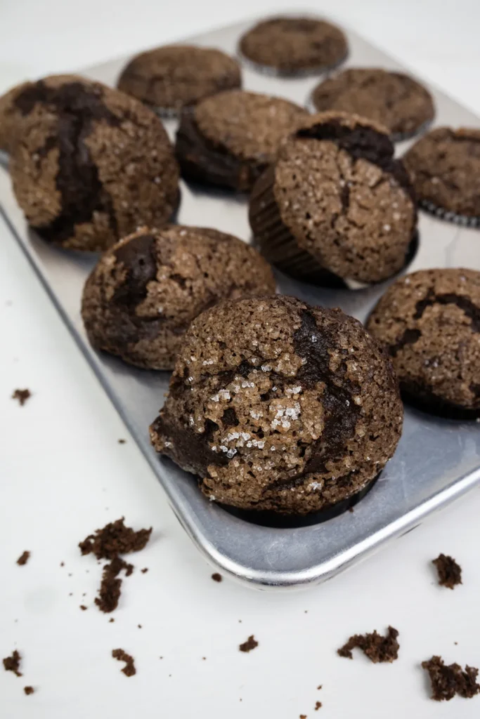 Baked sourdough double chocolate banana muffins in a muffin pan, with a few crumbs in the foreground.