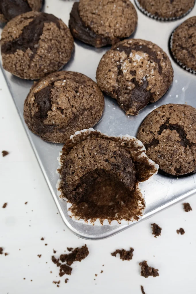 Sourdough double chocolate banana muffins in a muffin pan. One muffin has had a bite taken out of it.