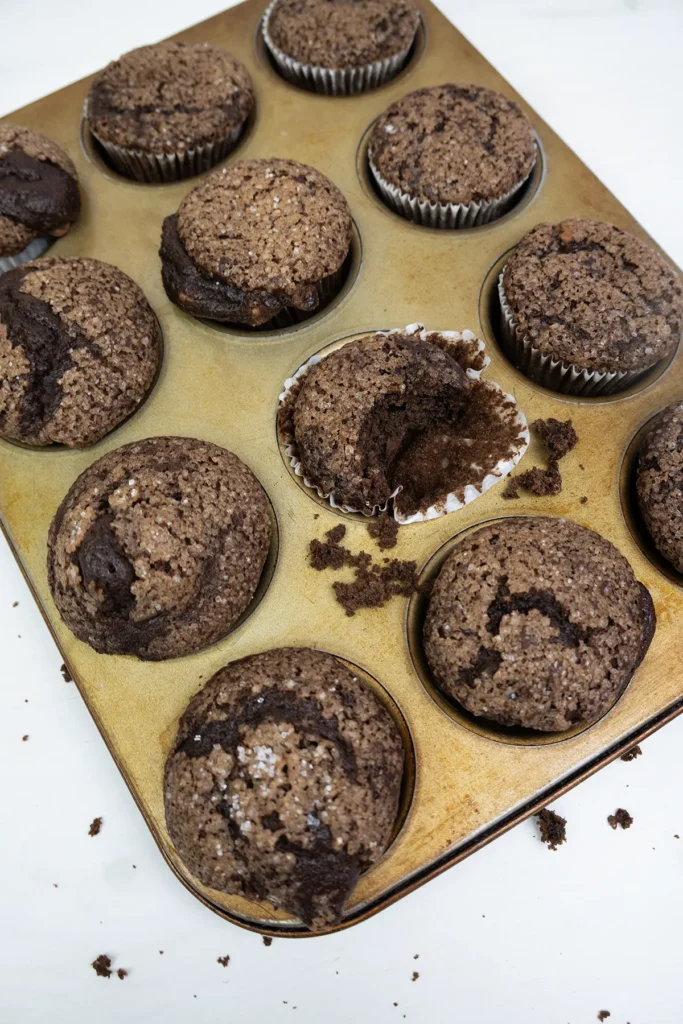 Overhead shot of sourdough double chocolate banana muffins in a muffin tray. One muffin has a bite taken out of it.