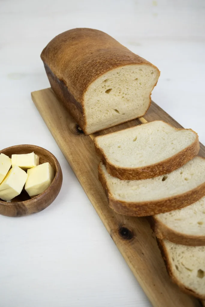 Sliced sourdough sandwich bread on a wooden board, with pieces of butter in a wooden bowl in the backgroud.