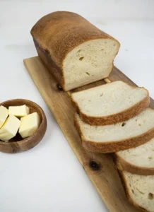 Slices of sourdough sandwich bread on a wooden board. There are cubes of butter in a wooden bowl in the background.