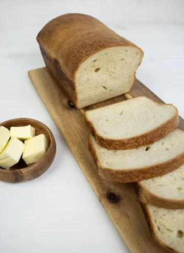 Slices of sourdough sandwich bread on a wooden board. There are cubes of butter in a wooden bowl in the background.