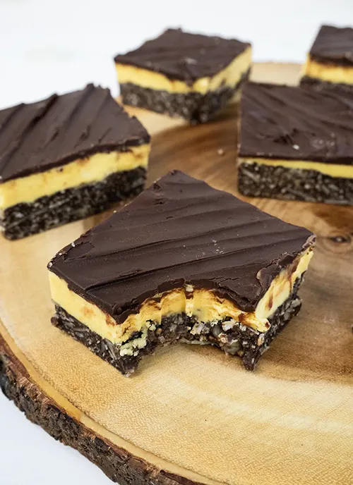 A wooden plate with scattered Nanaimo Bars. The one in the foreground has a bite taken out of it.