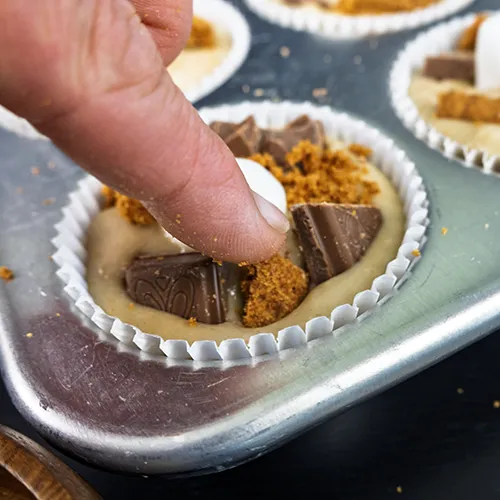Pressing chopped chocolate and graham crackers or speculoos cookies around the marshmallow of a sourdough banana s'mores muffin ready to be baked.