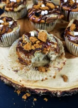 Sourdough banana s'mores muffins arranged on a wooden plate. A muffin in the foreground is unwrapped and has a bite taken out of it.