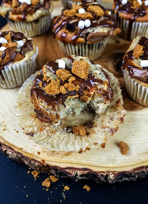 Sourdough banana s'mores muffins arranged on a wooden plate. A muffin in the foreground is unwrapped and has a bite taken out of it.