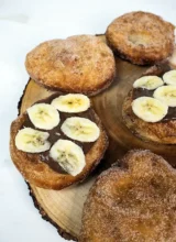 Mini sourdough Beaver Tails arranged on a wooden plate and coated with cinnamon sugar. Two of the Beaver Tails are topped with Nutella and banana slices.