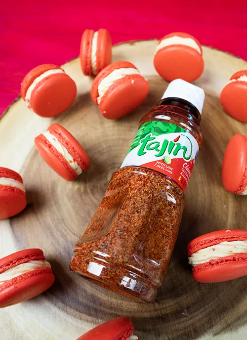 Strawberry Mango Tajín macarons scattered on a wooden platter, with a bottle of Tajín seasoning.