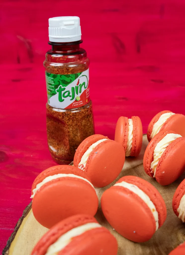 Strawberry Mango Tajín macarons scattered on a wooden platter, with a bottle of Tajín seasoning.