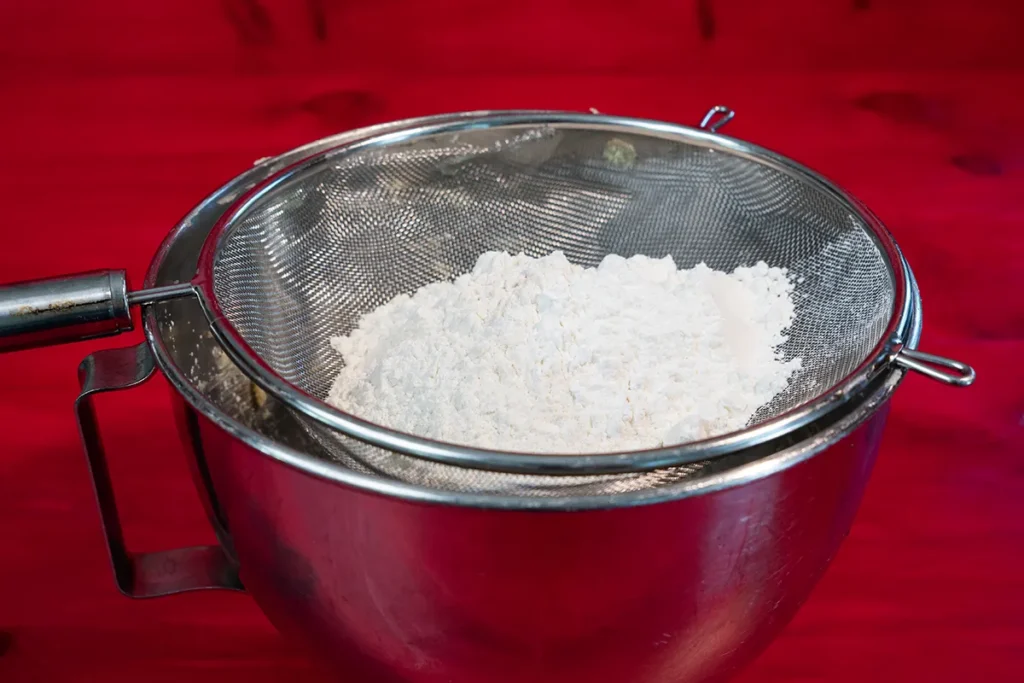 A sieve with all-purpose flour, cornstarch, and salt over a stand mixer bowl.