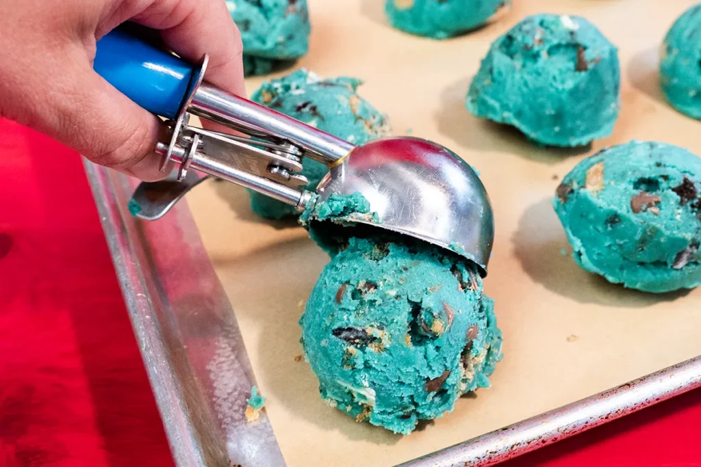Scooping blue cookie monster cookie dough onto a lined baking sheet.