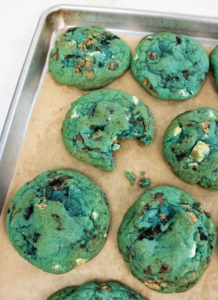 Blue cookie monster cookies arranged on a lined baking sheet. One has a bite taken out of it.