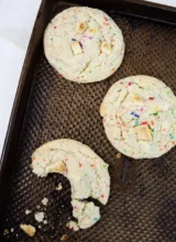 Three funfetti cake mix cookies arranged on a baking sheet. One cookie has a bite taken out of it.