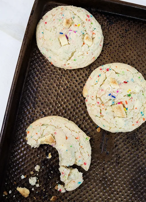 Three funfetti cake mix cookies arranged on a baking sheet. One cookie has a bite taken out of it.