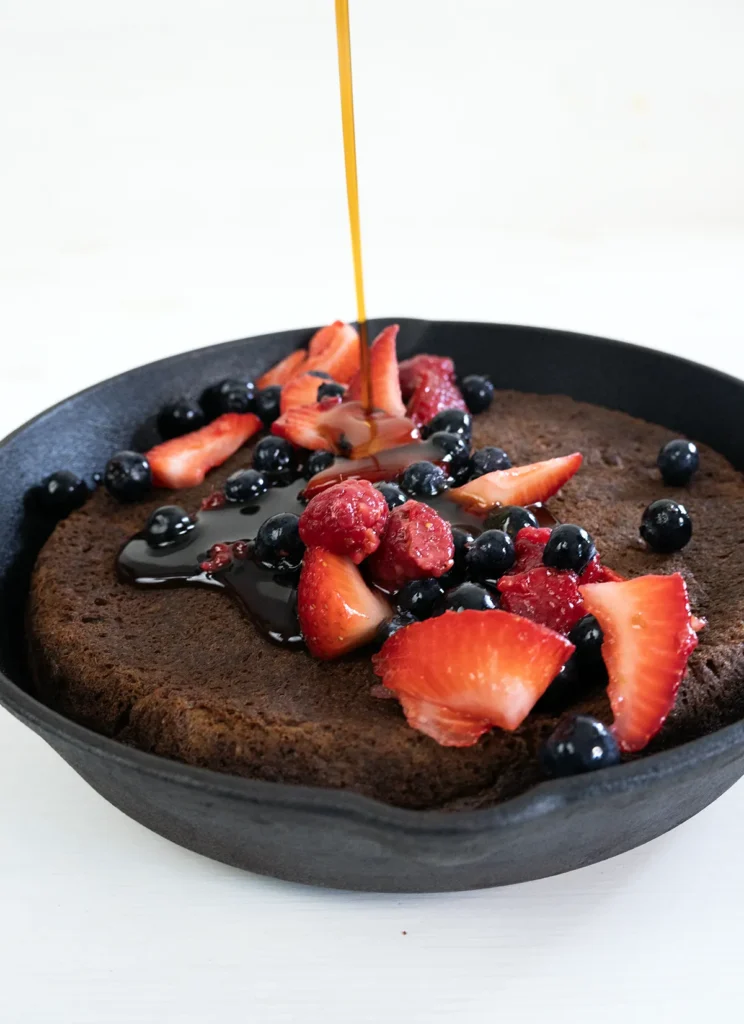 Pouring maple syrup over a giant skillet pancake topped with fresh berries.