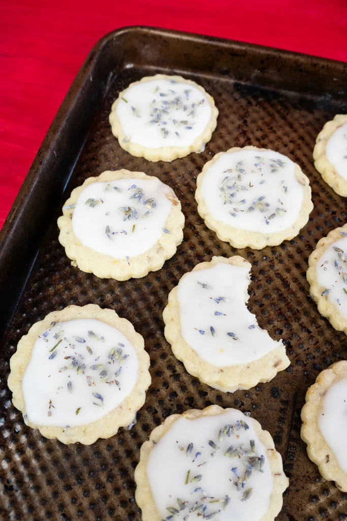 Glazed lemon lavender shortbread cookies on an old baking sheet. One cookie has a bite taken out of it.