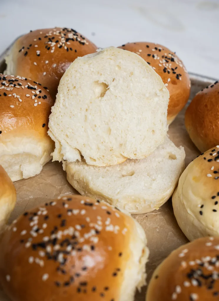 Baking sheet full of homemade burger buns topped with black and white sesame seeds. One bun is cut open, showing its crumb.