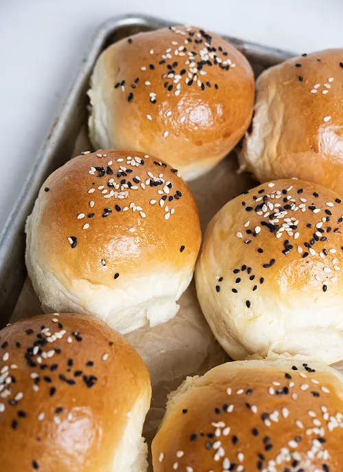Baking sheet full of homemade burger buns topped with black and white sesame seeds.